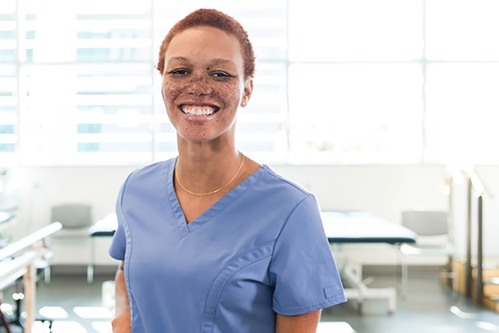A Black woman in scrubs smiles at the camera in a therapy facility.