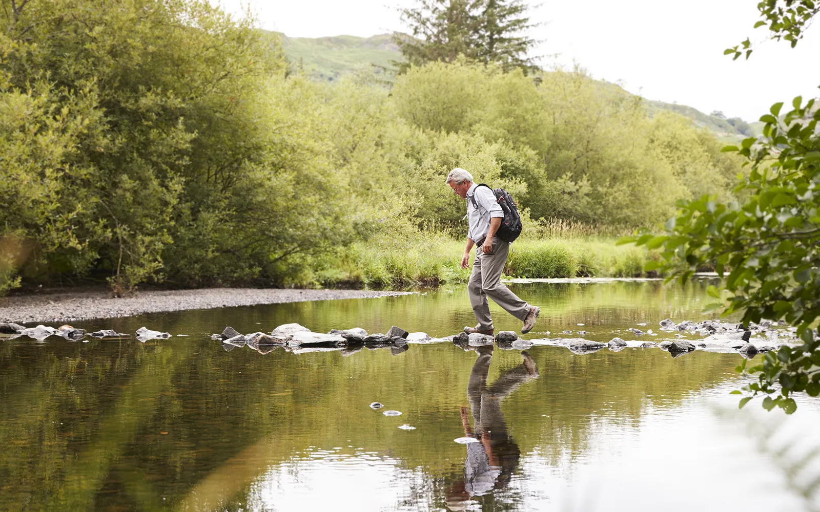 An elderly man steps on stones to cross a river.